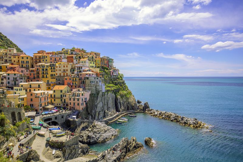 Pemandangan laut dari desa Manarola, salah satu dari lima kota di wilayah Cinque Terre di pesisir Italia. Pemandangan ini menangkap rumah-rumah berwarna-warni di tepi tebing di provinsi La Spezia, Liguria, dengan latar belakang hari musim panas yang cerah dengan langit biru dan awan putih. Manarola dikenal karena keindahan pemandangan dan arsitektur tradisionalnya, menjadi tujuan wisata terkenal yang mew exemplifies pesona khas Riviera Italia.