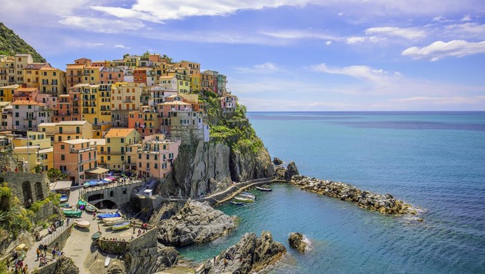 Pemandangan laut dari desa Manarola, salah satu dari lima kota di wilayah Cinque Terre di pesisir Italia. Pemandangan ini menangkap rumah-rumah berwarna-warni di tepi tebing di provinsi La Spezia, Liguria, dengan latar belakang hari musim panas yang cerah dengan langit biru dan awan putih. Manarola dikenal karena keindahan pemandangan dan arsitektur tradisionalnya, menjadi tujuan wisata terkenal yang mew exemplifies pesona khas Riviera Italia.