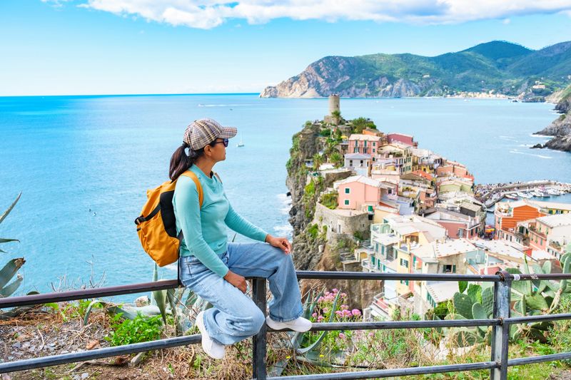 Seorang wisatawan wanita duduk di pagar pembatas, memandang garis pantai Cinque Terre yang menakjubkan. Rumah-rumah berwarna cerah yang bertengger di tebing dan laut yang berkilauan menciptakan latar belakang yang indah, Vernazza, Italia.