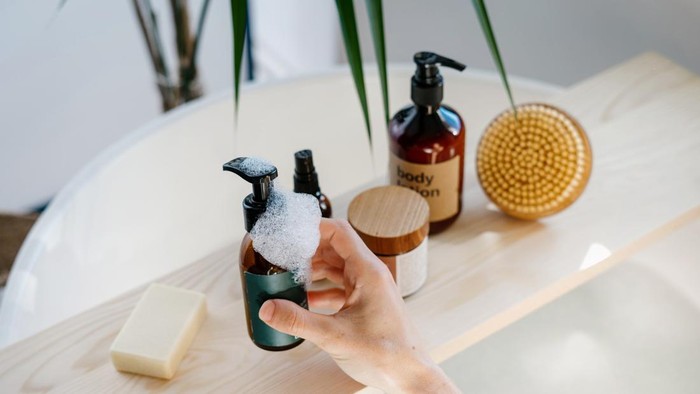 High angle view of woman hand choose cosmetics product for everyday skincare routine. Female hold glass bottle with dispenser over wooden shelf in bathroom. Self care, moisturizing, pampering concept
