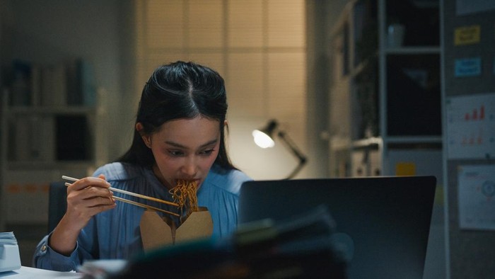 Overworked Asian businesswoman eating instant noodles while searching through paperwork late in busy office at night. Representing overtime, tight deadlines and the need to work through dinner at the desk.
