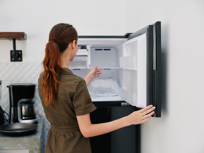 A woman in the kitchen of her home opened an empty freezer with ice inside, home refrigerator, defrosted, view from the back. High quality photo
