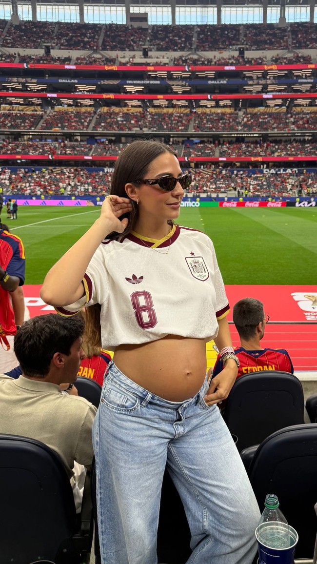 Rosa Pereira, istri dari gelandang Spanyol Fabián Ruiz, yang tengah hamil anak pertama mereka, bergaya memakai jersey Spanyol yang dibuat menjadi crop top sehingga memamerkan perutnya. Rosa berpose dengan baby bump dengan latar belakang Dallas Stadium tempat pertandingan Spanyol versus Portugal. Foto: Dok. Instagram