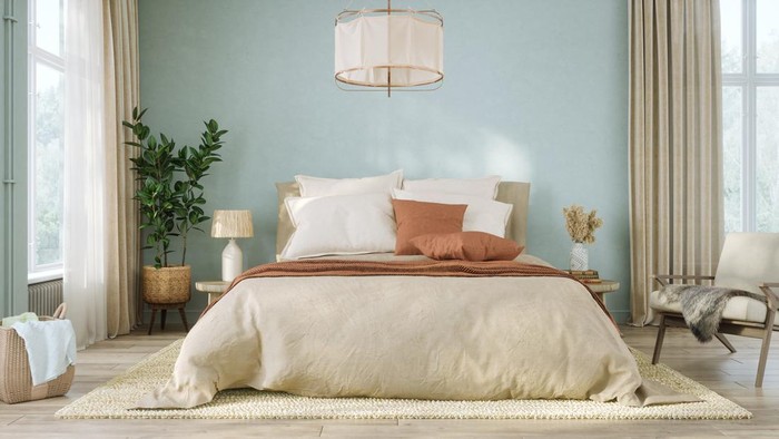 Contemporary bedroom features soft blue-green walls paired with beige linen bedding and rust-colored throw pillows. Wooden flooring, a textured area rug, and a large potted plant complement the natural aesthetic.