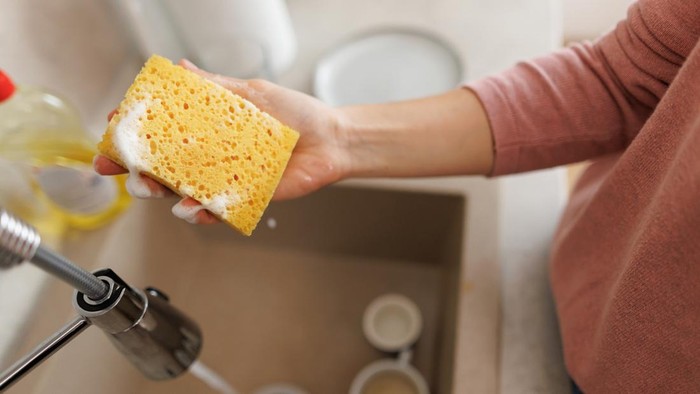 Woman holding a soapy sponge above a kitchen sink, preparing for dishwashing. Dish soap and dirty cups clutter the background, highlighting daily cleaning routines in a home setting