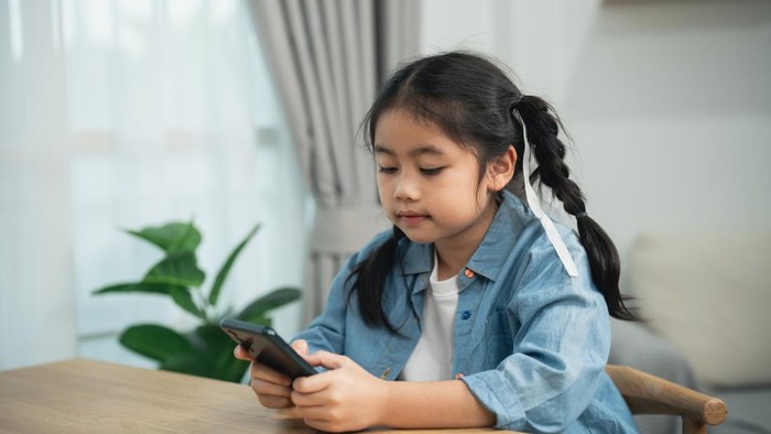 Asian girl using smartphone sitting at a table at home. Cute kid watching the screen, playing games, or learning online. Digital technology and childhood lifestyle concept.