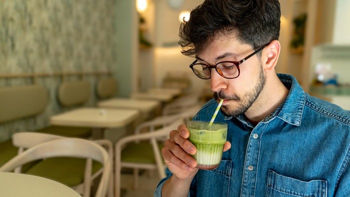 Young man smiling and holding a glass of green smoothie with a straw, sitting at a table in a restaurant