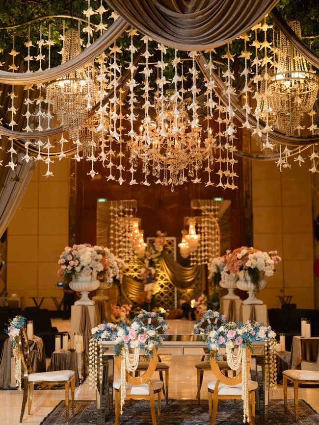 Dekorasi pernikahan Ayu dan Ruwadi tampak sangat megah dan elegan dengan langit-langit yang dihiasi bunga ronce melati dan lilin-lilin kristal yang mewah. Area akad nikah ditata dengan estetis menggunakan meja cermin yang memantulkan keindahan dekorasi, serta kursi beraksen emas dengan hiasan bunga bernuansa lembut. Foto: Dok. Instagram @ayupratiwi.