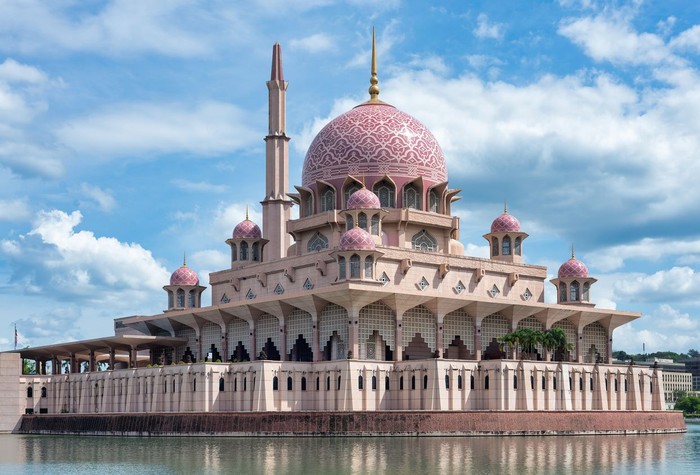 Putra Mosque, Putrajaya, Malaysia. Stunning pink mosque with a grand dome and minaret, reflected in the tranquil water under a bright blue sky with white clouds.
