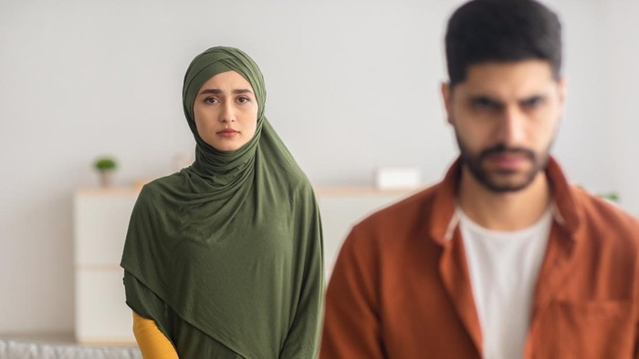 Unhappy Muslim Wife Standing Behind Husbands Back Looking At Camera In Fear At Home. Domestic Abuse Issue, Relationship With Abuser Concept. Selective Focus On Scared Woman