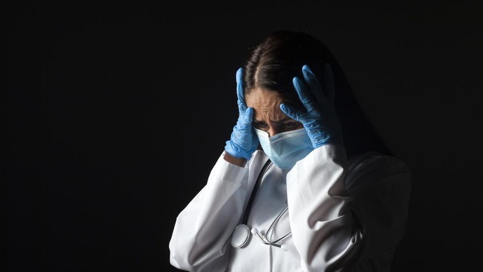 Doctor in disposable protective mask against coronavirus infection covid-19 holds head from fatigue isolated on black background