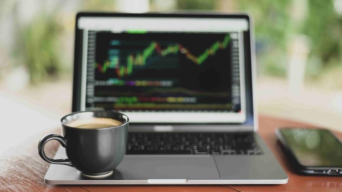 Ilustrasi grafik saham dalam latar belakang hitam pada layar laptop dan secangkir kopi