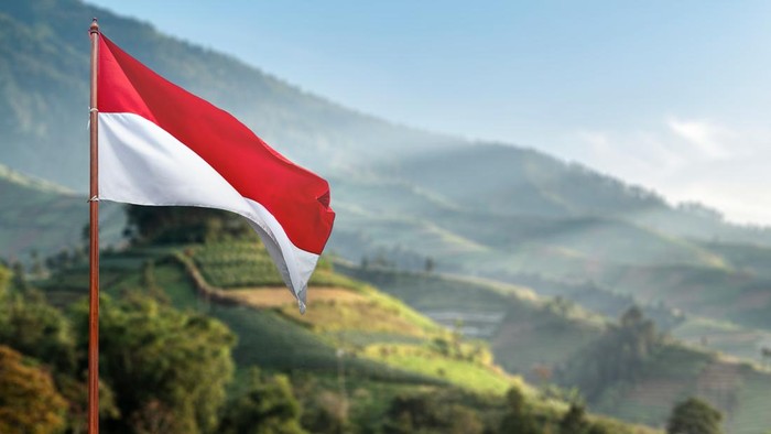 The red and white national flag of Indonesia waving on a flagpole against a beautiful mountain landscape and green terraced hills. Hari Kemerdekaan, Merdeka, and Dirgahayu Indonesia on 17 Agustus