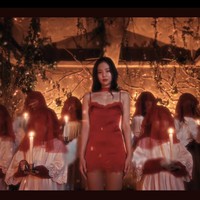 Netizen semakin geger usai melihat adegan Jennie BLACKPINK berbalut gaun satin merah. Berdiri di tengah kumpulan anak-anak yang memakai tudung kepala, kostum putih menyerupai busana misdinar gereja, sambil memegang lilin. Foto: dok. YouTube JENNIE