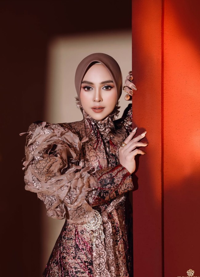 Bagian paling mencolok adalah lengan kiri yang dibuat sangat bervolume dengan susunan ruffle atau layer kain bertumpuk. Atasannya juga memiliki model kerah tinggi dengan aksen kain yang tampak seperti ruffle atau drapery di area leher dan dada. Foto: Instagram/@riaricis1795