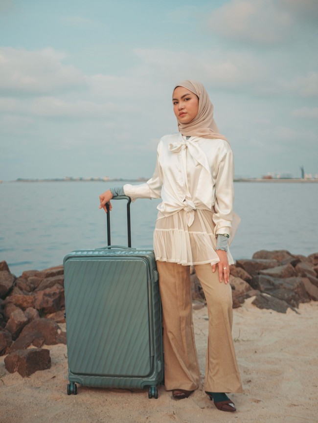 Xaviera Putri memakai pashmina segi empat nude bernuansa earth tone yang dililit rapi ke belakang. Outfit memadukan blouse putih aksen pita dengan luaran tulle transparan serta celana bahan high-waist berwarna krem netral yang elegan. Foto: Dok. Instagram @xavieraaputri.