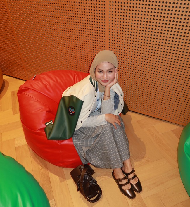 Xaviera Putri memakai pashmina cokelat muda dengan penataan melingkar rapi di kepala. Tampilan outfit bergaya preppy casual memadukan kemeja, rok plisket kotak-kotak, serta jaket varsity kombinasi krem-hijau. Foto: Dok. Instagram @xavieraaputri.