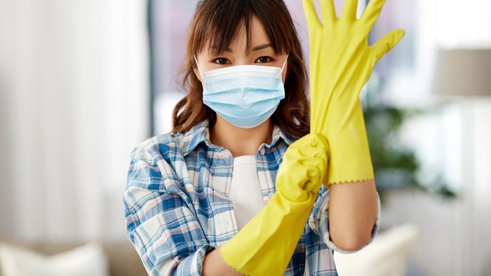 cleaning, health and hygiene concept - asian woman wearing protective medical mask for protection from virus disease and putting rubber gloves on at home