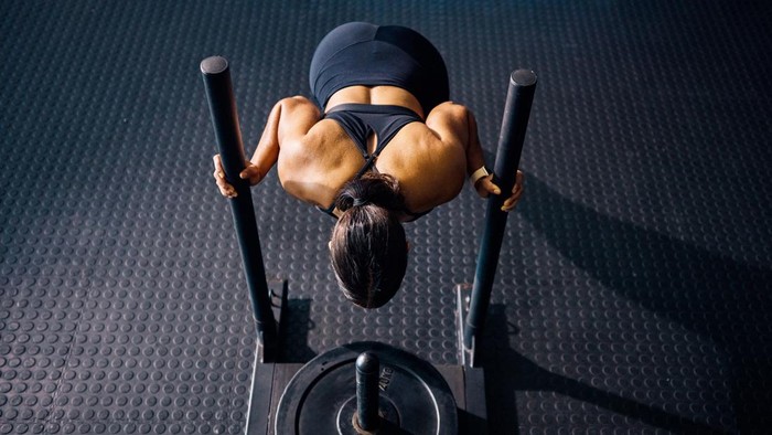 Female athlete pushing a weighted sled in a gym. Top view of a woman performing strength training exercise on a black gym floor. Hyrox Training Concept.