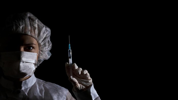 Female doctor (or nurse) in protection face mask, medical cap and glove holds syringe with vaccine in her hand on black background. Half face in shadow, dramatic emotion. Concept of virus protection, prevention of epidemics, treatment of diseases. Ba