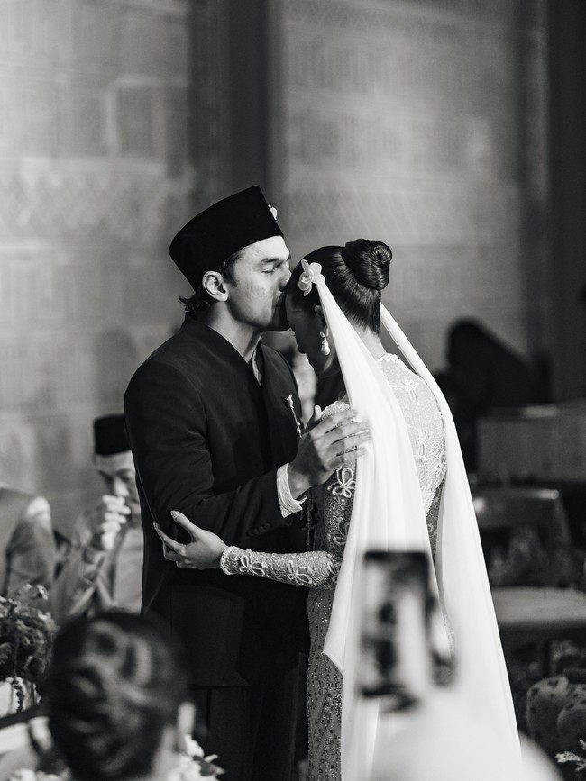 Sementara itu, Givina tampil dalam gaun pengantin putih dengan siluet fitted. Busananya memiliki detail transparan pada bagian lengan yang dihiasi motif floral serta embellishment berkilau. Bagian bodice hingga rok dipenuhi detail payet dan kristal yang membentuk pola memanjang. Siluetnya mengikuti bentuk tubuh sebelum melebar di bagian bawah, memberikan kesan seperti mermaid gown.  Foto: Instagram @caesarmario