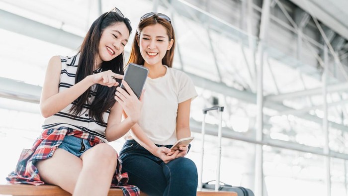 Two happy Asian girls using smartphone checking flight or online check-in at airport together, with luggage. Air travel, summer holiday, or mobile phone application technology concept