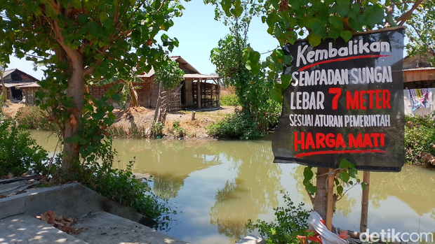 Baliho protes yang dipasang di tepi aliran sungai menuju tambak, akses melewati tembok bolong. Baliho protes yang dipasang di tepi aliran sungai menuju tambak, akses melewati tembok bolong.
