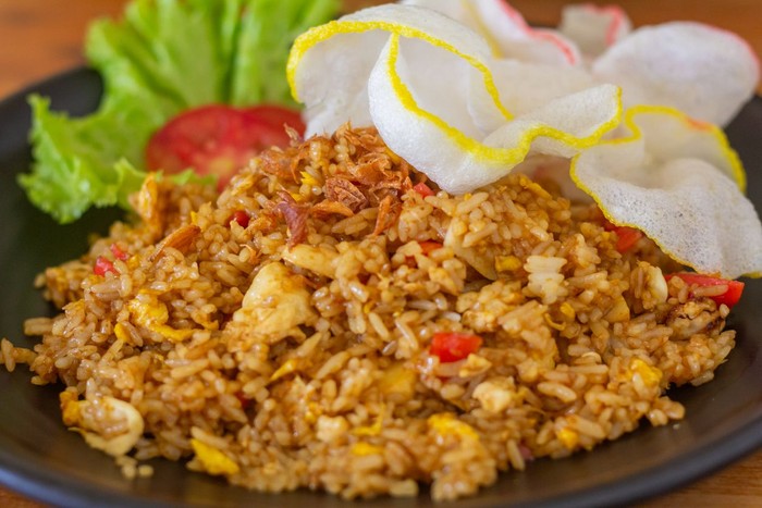 Delicious Indonesian fried rice served on a black plate with crispy prawn crackers, lettuce, tomato slice, and fried chalots topping.