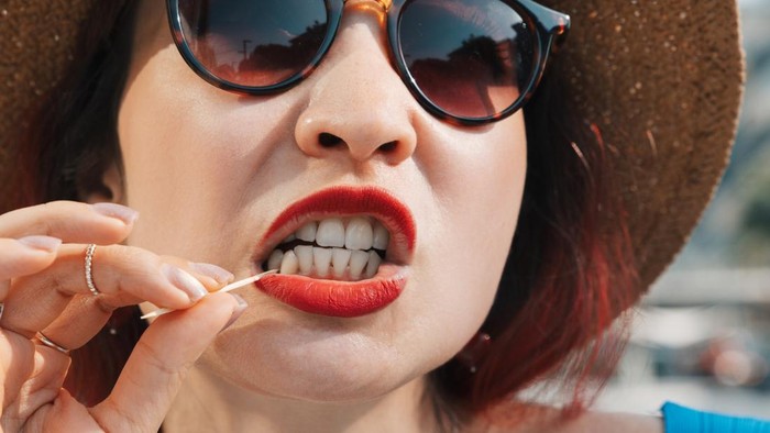 Woman in sunglasses and a straw hat cleaning her white teeth with a toothpick, enjoying personal oral hygiene on a sunny day