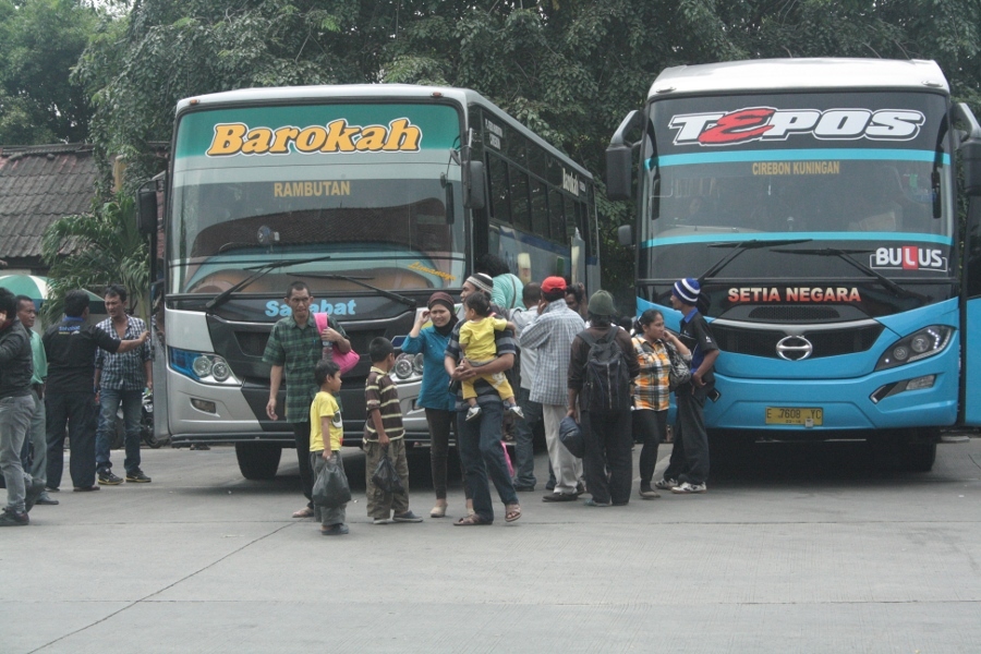 Sebuah bus pariwisata berwarna cerah melintas di jalanan Kota Cirebon dengan latar belakang bangunan bersejarah dan pepohonan hijau, menunjukkan suasana kota yang ramai namun tenang.
