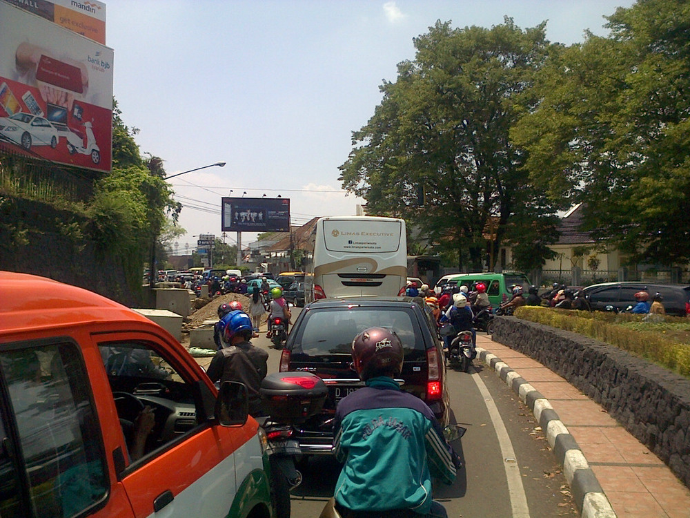 Pemandangan jalan raya perkotaan yang padat dengan lalu lintas mobil dan motor yang merayap perlahan, menunjukkan kemacetan parah dengan banyak kendaraan yang berhenti di beberapa lajur jalan di kota Bandung pada siang hari.