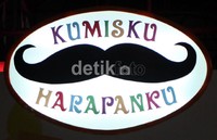 Dibawah logo yang bertuliskan Kumisku Harapanku ada gambar Monas, gedung bertingkat, jembatan layang, mobil dan kereta.