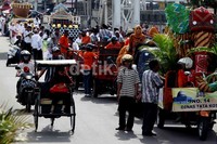 Sebuah becak yang ditumpangi oleh seorang ibu, melintas di sebelah arak-arakan kendaraan hias.