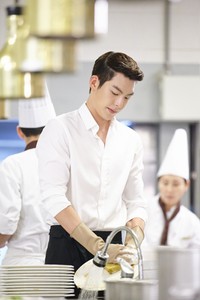 Dalam foto tersebut, Woobin mengenakan kemeja putih sambil mencuci piring di sebuah dapur. (Hwa and Dam Pictures)