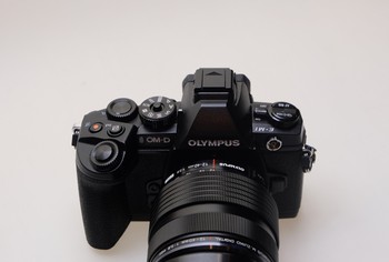 Di bagian atas, ada mode dial hingga dua control dial, satu di depan dan satu di belakang. Asyiknya, Olympus menyediakan satu tombol switch yang terletak di samping EVF (Electronic Viewfinder) untuk berganti konfigurasi tersebut. Ada juga tombol kunci di tengah mode dial untuk mencegah pengguna tak sengaja salah berpindah mode.     