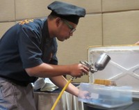 Chocolate Tonka Bar disemprot dengan mesin kompressor yang berisi lemak cokelat dan pewarna coeklat untuk mempercantik sajian cake.