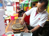 Koh Asan sedang menaburkan susu kental manis, meisjes, dan kacang giling yang ia buat sendiri.