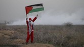 Seorang demonstran berpakaian seperti Sinterklas sambil memegang bendera Palestina ketika tentara Israel menyemprotkan gas air mata dalam demonstrasi memprotes keputusan Presiden AS Donald Trump yang mengakui Yerusalem sebagai ibu kota Israel di timur Gaza, sekat perbatasan dengan Israel, Selasa (19/12). (Reuters /Mohammed Salem)