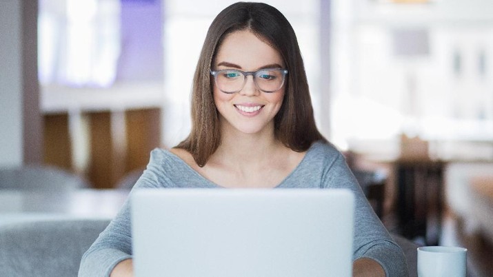 Portrait of cheerful Caucasian female student wearing eyeglasses using laptop in cafe for studies or networking and smiling