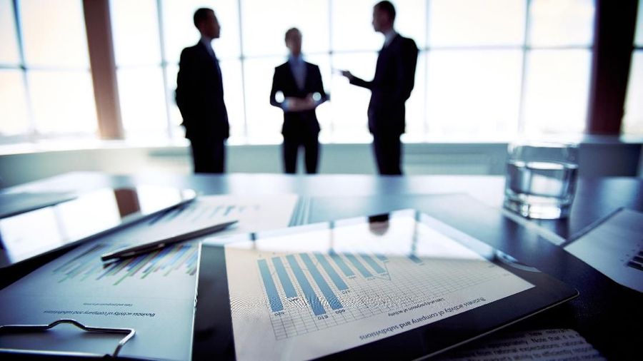 Close-shot of a tablet computer displaying financial data, three businessmen standing in the background
