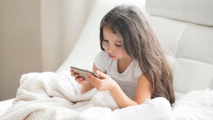 Little girl playing games or using app on tablet sitting on white coach in living room. Beautiful kid girl sitting on cozy white home sofa with blanket and looking at smartphone screen