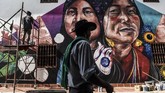 Warga yang melintas menyempatkan diri memperhatikan seorang artis mural yang sedang menggambar di perancah, di First International Mural Festival for Peace di San Carlos, Kolombia. (Photo by JOAQUIN SARMIENTO / AFP)