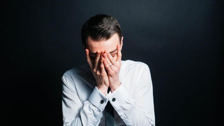 Color portrait of a young man in a shirt and black tie, upset, hands on his face, against plain studio background.