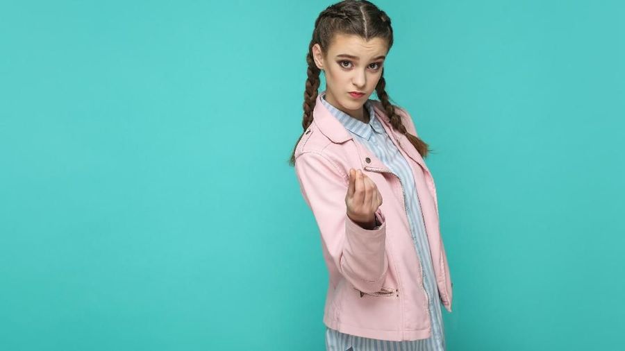 money gesture. portrait of beautiful cute girl standing with makeup and brown pigtail hairstyle in striped light blue shirt pink jacket. indoor, studio shot isolated on blue or green background.