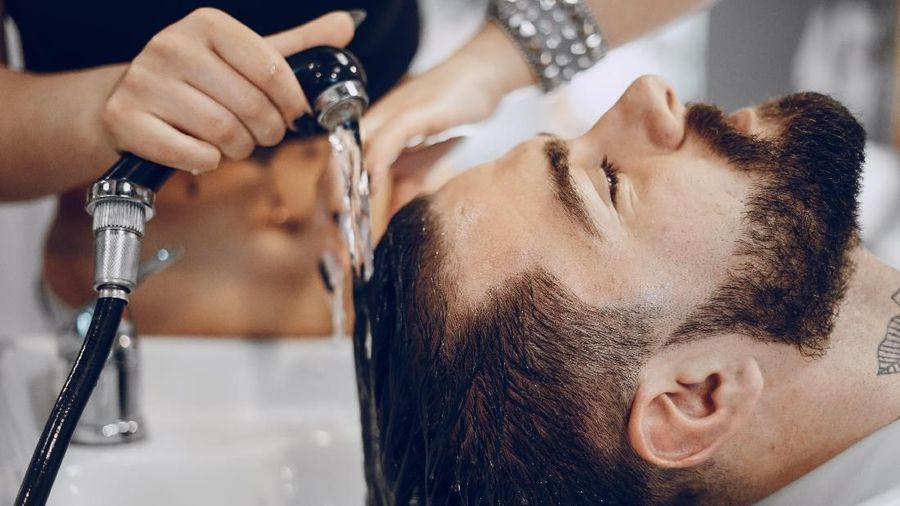 handsome young bearded guy sitting in an armchair in a beauty salon and the girl near him washes his head under the jet of water