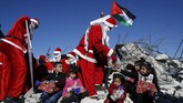 Warga Palestina mengenakan kostum Natal dan membagikan hadiah kepada anak-anak yang duduk di atas puing sebuah rumah yang dihancurkan Israel pada Senin (23/12). (Photo by Musa Al SHAER / AFP)