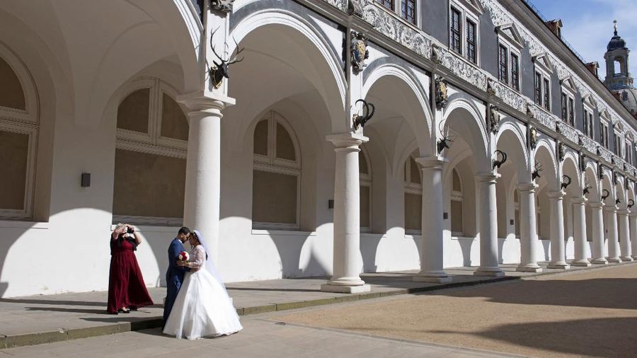The just married couple Laila, right, and Boualem Bellil are photographed by Laila's mother, left, in the Stall Courtyard, a most frequented tourist spot, in Dresden, eastern Germany, Thursday, March 26, 2020. They celebrate today without wedding guests because of the government regulations and want to make up for the party next year. In order to slow down the spread of the coronavirus, the German government has considerably restricted public life and asked the citizens to stay at home. The new coronavirus causes mild or moderate symptoms for most people, but for some, especially older adults and people with existing health problems, it can cause more severe illness or death. (AP Photo/Jens Meyer)
