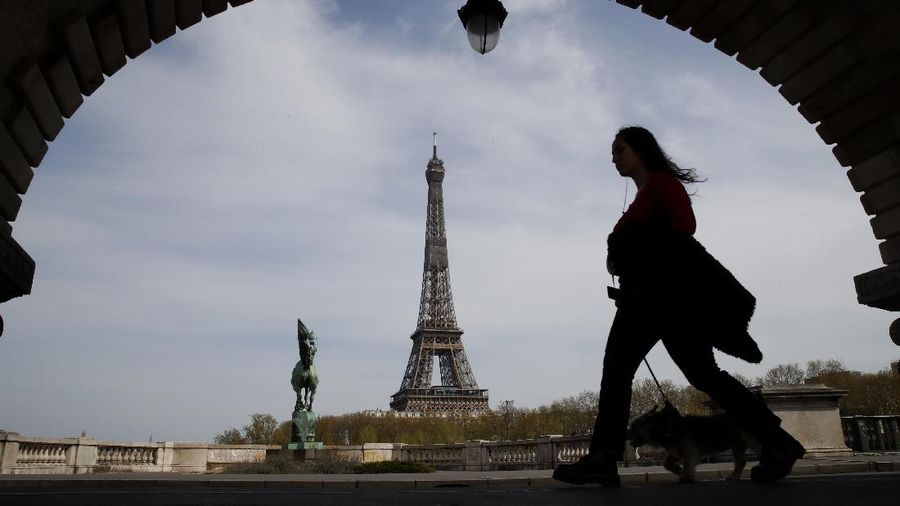 A woman walks her dog on a Paris bridge, with the Eiffel tower seen in background, during a nationwide confinement to counter the COVID-19, Tuesday, April 7, 2020. The new coronavirus causes mild or moderate symptoms for most people, but for some, especially older adults and people with existing health problems, it can cause more severe illness or death. (AP Photo/Christophe Ena)