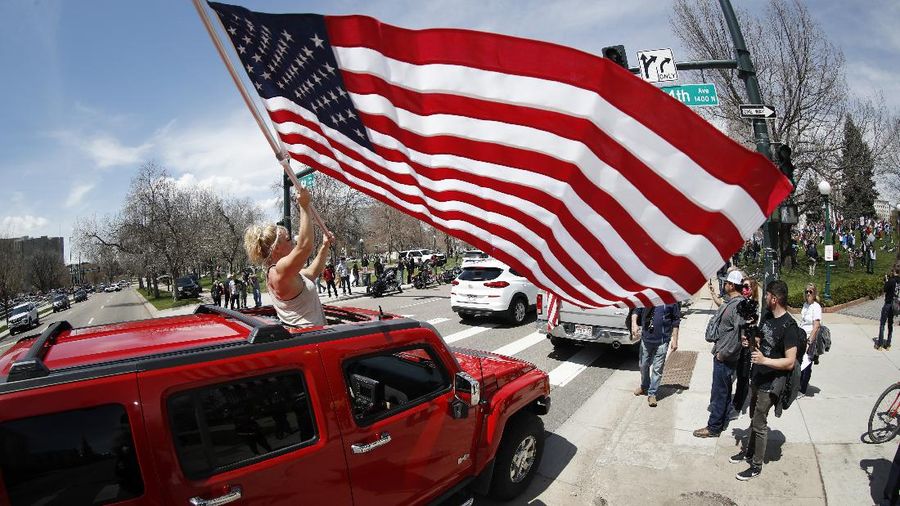 A protester stands out of the sunroof of a sports-utility vehicle to wave an American flag during a car protest against the stay-at-home order issued by Colorado Governor Jared Polis to stem the spread of the new coronavirus Sunday, April 19, 2020, in Denver. (AP Photo/David Zalubowski)