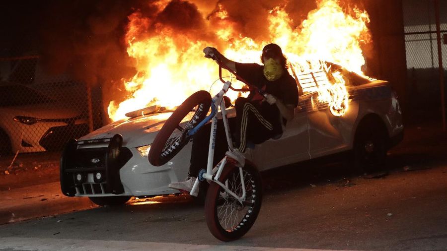 A protester on a bicycle rides past a burning police car during a demonstration next to the city of Miami Police Department, Saturday, May 30, 2020, downtown in Miami. Protests were held throughout the country over the death of George Floyd, a black man who was killed in police custody in Minneapolis on May 25. (AP Photo/Wilfredo Lee)